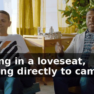 sitting in a loveseat, talking directly to camera.